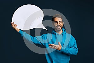 Cheerful young man in eyeglasses and blue hoodie holding blank speech bubble