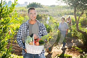 Cheerful young man holding basket of fresh vegetables in garden