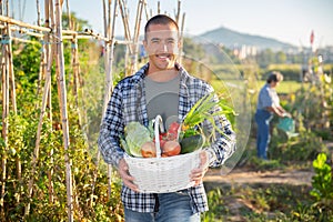 Cheerful young man holding basket of fresh vegetables in garden