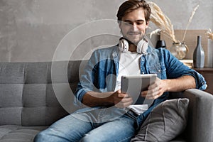 Cheerful young man holding digital tablet and looking relaxed while lying on the couch at home