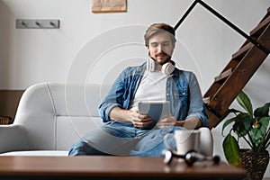 Cheerful young man holding digital tablet and looking relaxed while lying on the couch at home