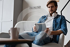 Cheerful young man holding digital tablet and looking relaxed while lying on the couch at home