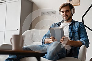 Cheerful young man holding digital tablet and looking relaxed while lying on the couch at home