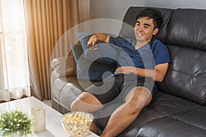 Cheerful young man holding remote control and watching TV while