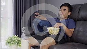 Cheerful young man holding remote control and watching TV while sitting on sofa