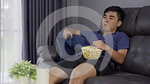 Cheerful young man holding remote control and watching TV while sitting on sofa