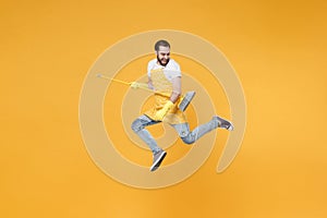 Cheerful young man househusband in apron rubber gloves hold broom like guitar while doing housework isolated on yellow