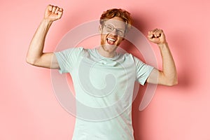 Cheerful young man with red hair looking happy, raising hands up in fist pumps gesture, celebrating success, feel like