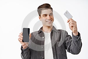 Cheerful young man shows smartphone screen, raise hand with credit card, paying online, shopping contactless in internet