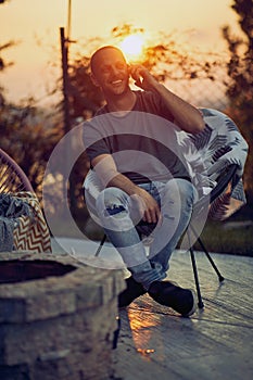 Cheerful young man sitting outdoors in a cozy chair on a patio, eith sunset in the background, talking on the phone, feeling