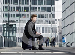 Cheerful young man sitting on suitcase looking at mobile phone