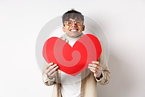 Cheerful young man smiling and looking thrilled at camera, showing big red heart cutout on Valentines day, making