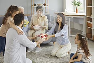 Cheerful young man surrounded by the whole family gives birthday presents to his happy wife.