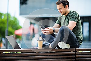 Cheerful young man using mobile phone while sitting at the city park, drinking coffee