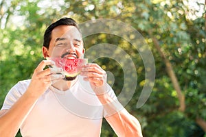 Cheerful young man in white shirt biting juicy slice of red watermelon outdoors in summer day. Enjoying life concept
