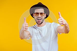 cheerful young man in white t-shirt, hat and sunglasses showing thumbs up and smiling at camera