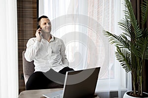 Cheerful Young Man Working Remotely From Home Office With Cellphone And Laptop, Sitting At Desk In Room, Copy Space.