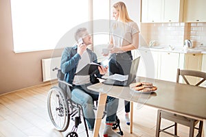 Cheerful young man with inclusiveness sit at table and talk on phone. He look at woman and smile. Holding opened folder