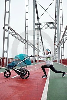 Cheerful young mom exercising on the bridge with a baby carriage, preparing for a run on a cloudy day in the city