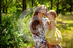 Cheerful young mother holds her little baby girl and smiles at the green park