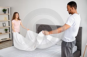 Cheerful young multinational couple making bed at home, holding blanket, doing housework together