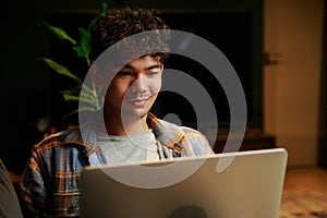 Cheerful young multiracial man wearing checkered shirt using laptop on sofa in house