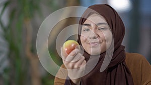 Cheerful young Muslim woman in hijab smelling organic healthful apple looking at camera and smiling. Portrait of happy