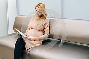 Cheerful young pregnant woman is sitting in hallway of hospital waiting to see gynecologist.