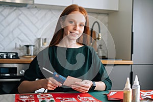 Cheerful young redhead woman gluing envelopes on board with gifts for children making Christmas advent calendar at home