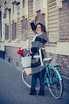 Cheerful young smiling woman is walking with bicycle and waving hand on city street