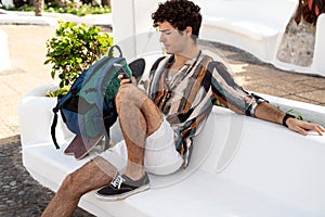 Cheerful young spanish man sitting outdoor and using mobile phone, scrolling through social media. Traveler guy talking online at