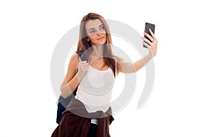 Cheerful young student girl with backpack makes selfie on her mobile phone isolated on white background. student years