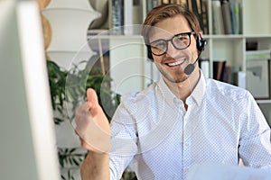 Cheerful young support phone male operator in headset, at workplace while using computer