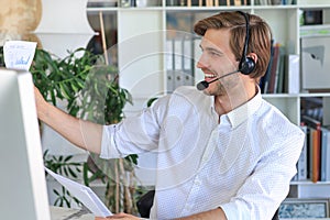 Cheerful young support phone male operator in headset, at workplace while using computer