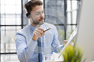 Cheerful young support phone male operator in headset, at workplace while using computer