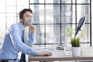 Cheerful young support phone male operator in headset, at workplace while using computer