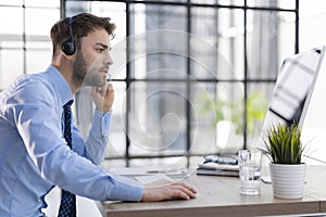 Cheerful young support phone male operator in headset, at workplace while using computer