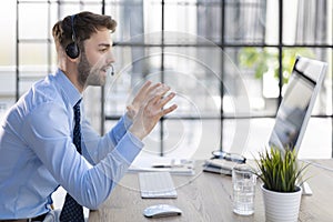 Cheerful young support phone male operator in headset, at workplace while using computer