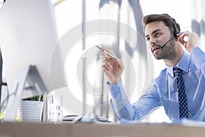 Cheerful young support phone male operator in headset, at workplace while using computer