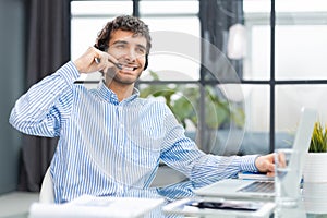 Cheerful young support phone male operator in headset, at workplace while using computer, help service and client