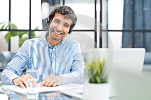 Cheerful young support phone male operator in headset, at workplace while using computer, help service and client