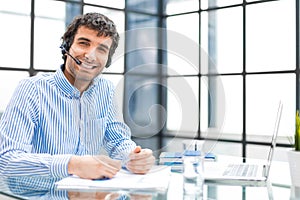 Cheerful young support phone male operator in headset, at workplace while using computer, help service and client