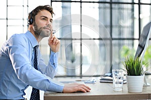 Cheerful young support phone male operator in headset, at workplace while using computer, help service and client