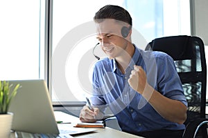 Cheerful young support phone male operator in headset, at workplace while using laptop