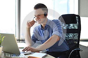 Cheerful young support phone male operator in headset, at workplace while using laptop