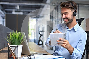 Cheerful young support phone male operator in headset, at workplace while using laptop