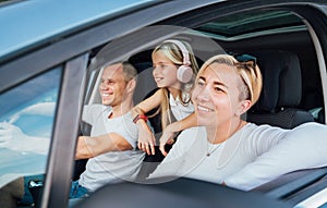 Cheerful young traditional family has a auto journey in modern car. Mother, father and daugher with headphones. Safety riding car