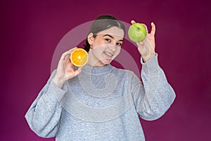 Cheerful young woman with apple and orange on purple background.