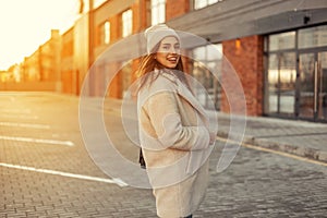 Cheerful young woman with a beautiful smile in a fashionable hat in a stylish coat stands in the parking on the background of a