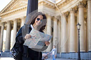Cheerful young woman in black, in sunglasses with a city paper map in hands, have a backpack, on cathedral background.